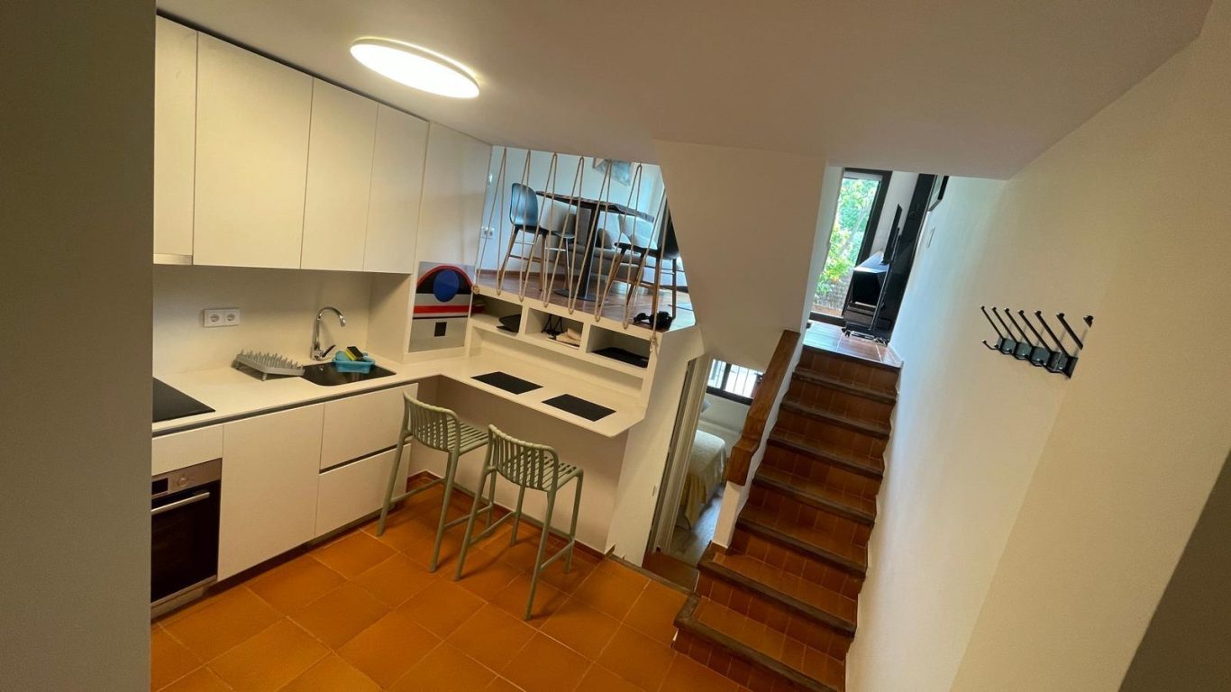 Cocina moderna con barra, escaleras de madera y acceso a planta superior iluminada.